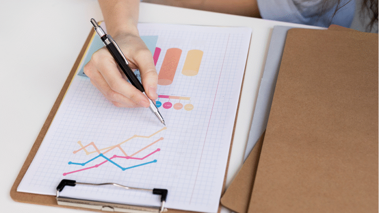 Hand holding a pen and drawing colorful line graphs on grid paper clipped to a clipboard.